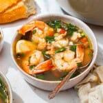 A close up of a bowl of seafood stew with a spoon. Next to this is a kitchen towel, another bowl of the stew, a bowl of lemon wedges, the loaf of crusty bread, and a bowl of black pepper.