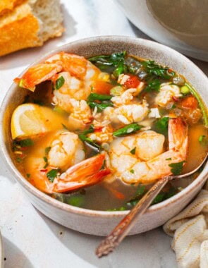 A close up of a bowl of seafood stew with a spoon. Next to this is a kitchen towel, another bowl of the stew, a bowl of lemon wedges, the loaf of crusty bread, and a bowl of black pepper.