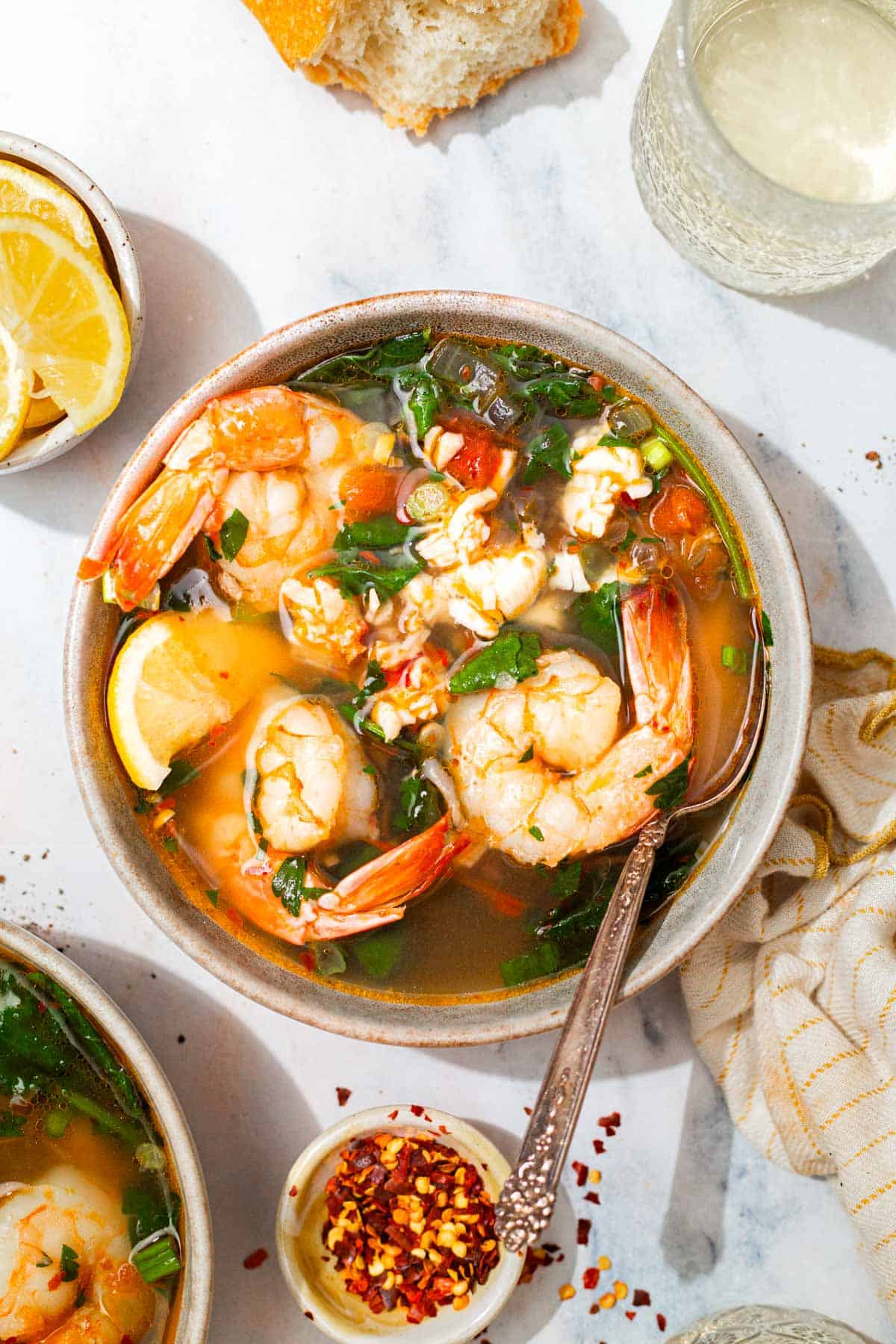 An overhead photo of a bowl of seafood stew with a spoon. Next to this is another bowl of the stew, a bowl of lemon wedges, a piece of crusty bread, a glass of white wine, a kitchen towel and a small bowl of red pepper flakes.