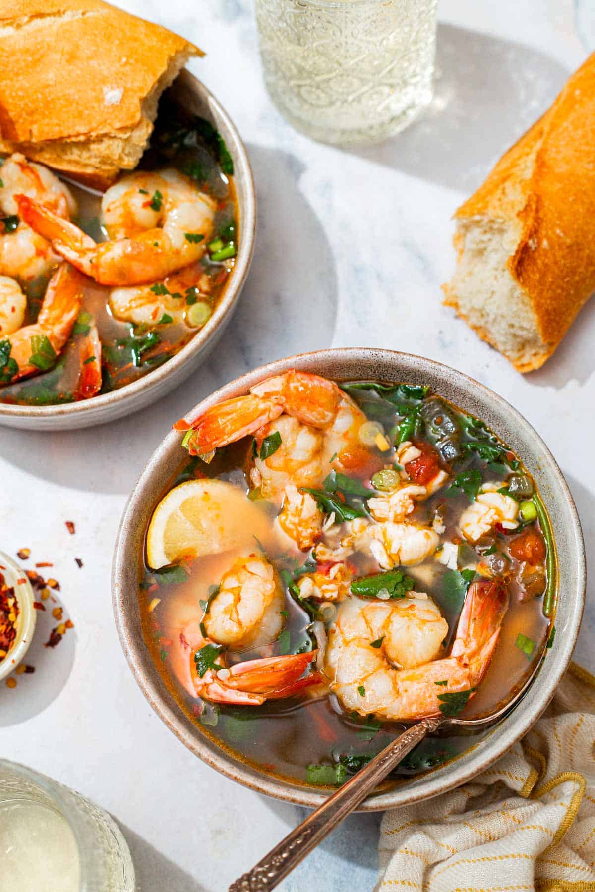 An overhead photo of a bowl of seafood stew with a spoon. Next to this is another bowl of the stew with a piece of crusty bread, the loaf of crusty bread, 2 glasses of white wine, a kitchen towel and a small bowl of red pepper flakes.