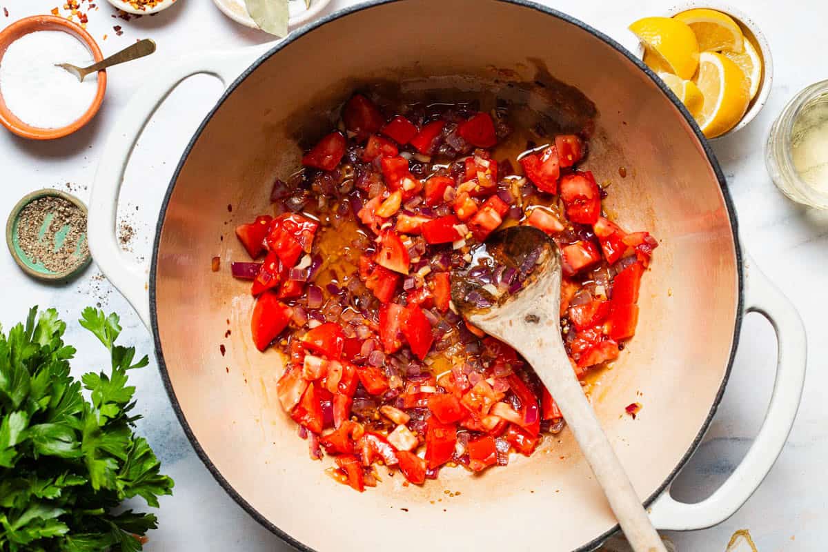 Seasoned and chopped red onions, garlic, and tomatoes being sauteed in a large pot with a wooden spoon. Surrounding this is a bunch of parsley, a cup of white wine, and bowls of black pepper, salt, and lemon wedges.