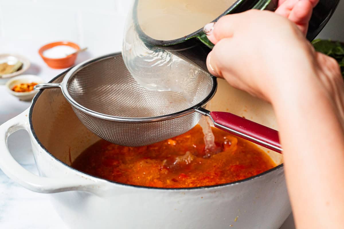 A close up of the lobster broth being poured through a mesh strainer into the pot with the sauteed vegetables.