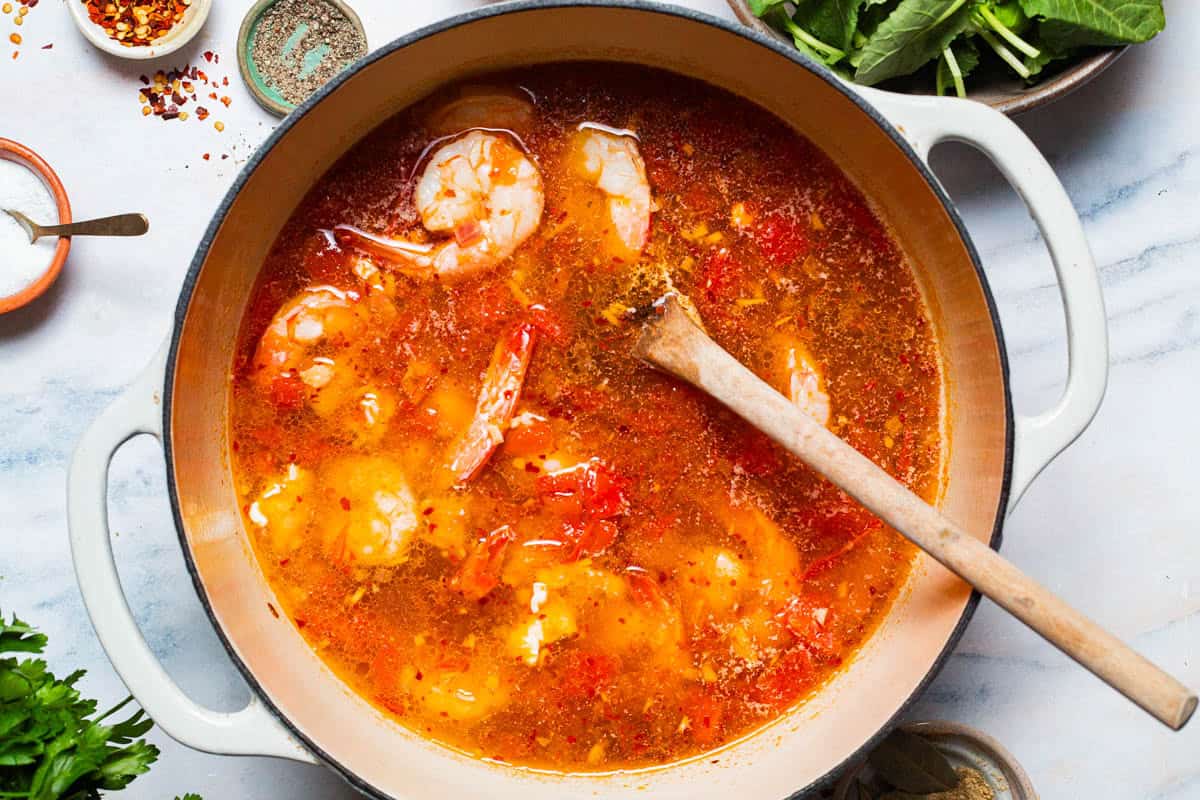 An overhead photo of the seafood stew in a large pot with a wooden spoon before the kale is added. Surrounding this are the various other ingredients for the stew.