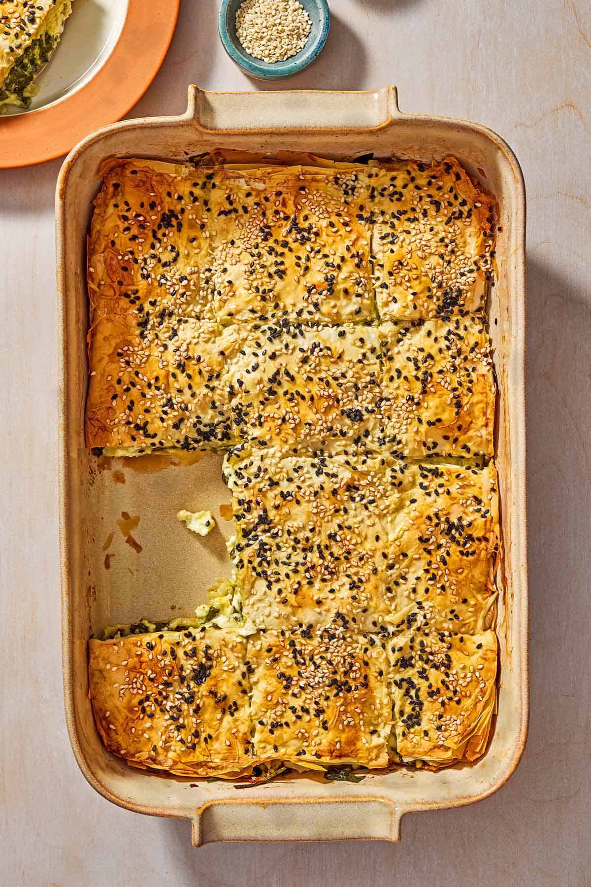 An overhead photo of sliced turkish borek in a baking dish with a piece removed. Next to his is a piece of the borek on a plate, and a small bowl of sesame seeds.