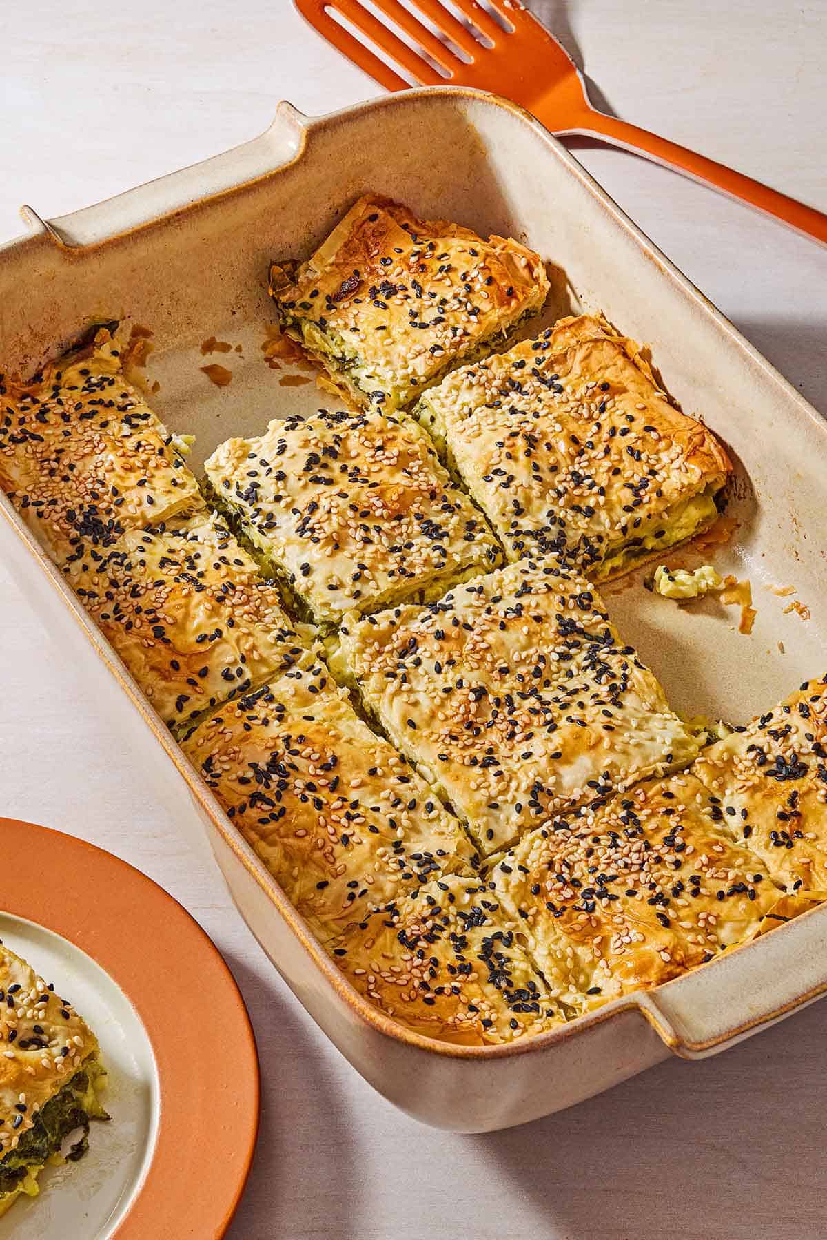 Sliced turkish borek in a baking dish with a few pieces removed. Next to this is a spatula and a piece of the borek on a plate.