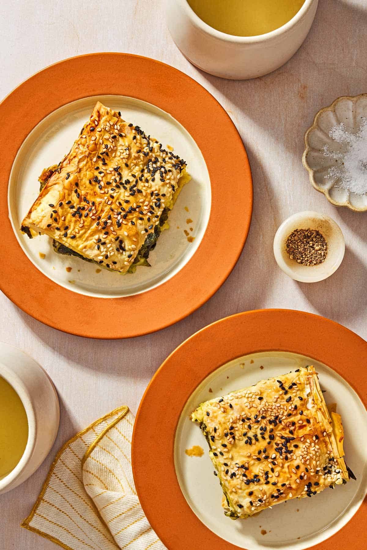 An overhead photo of 2 pieces of Turkish borek on plates. Next to these are cups of orange juice, bowls of salt and pepper and a cloth napkin.