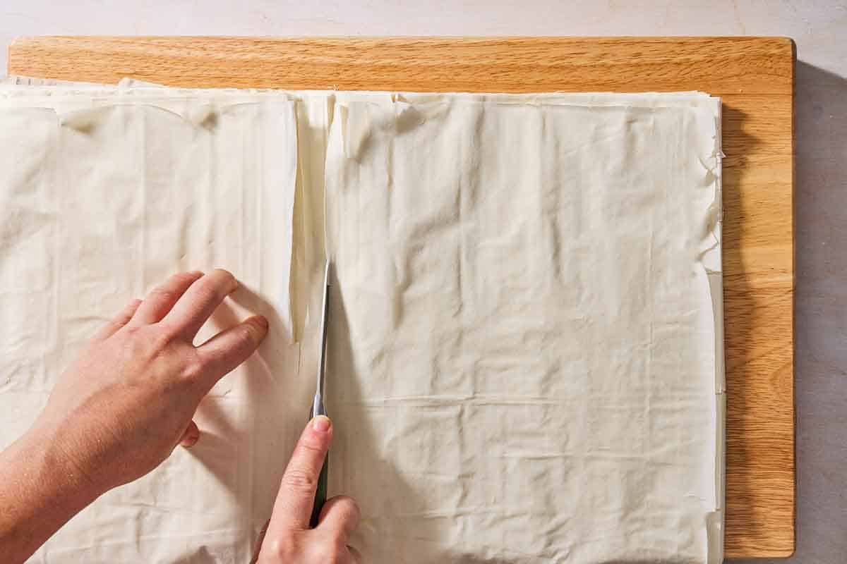 An over head photo of a stack of several sheets of phyllo dough being cut in half on a cutting board with a knife.