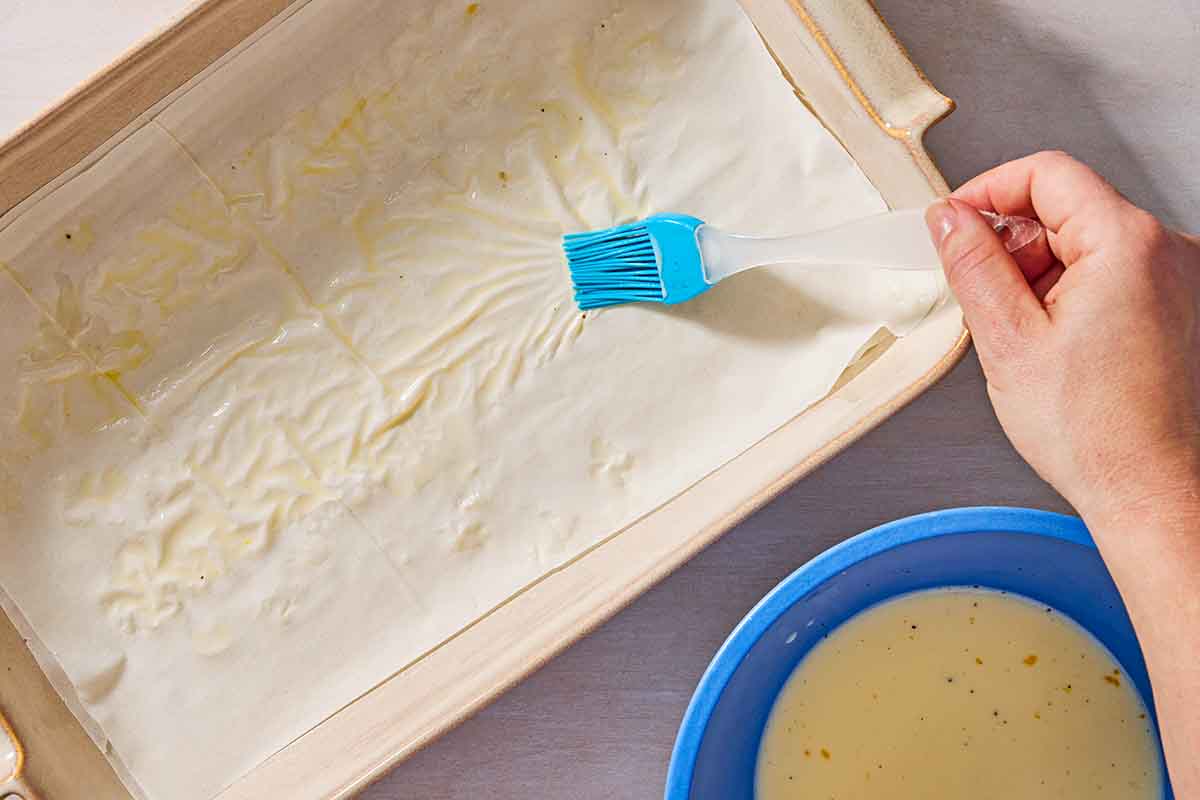 An overhead photo of the egg and milk mixture being brushed onto the first layers of phyllo dough in a baking dish. Next to this is a bowl of the milk and egg mixture.