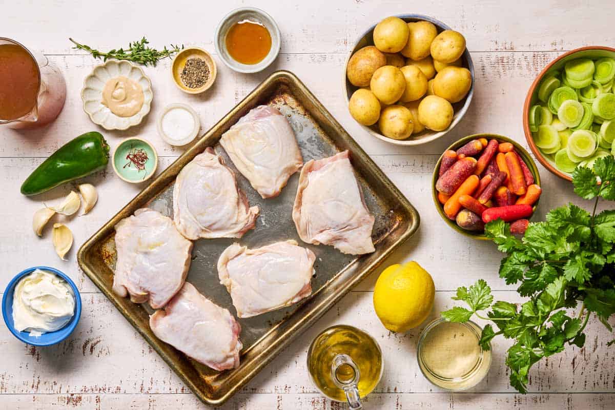 An overhead photo of the ingredients for creamy saffron chicken including bone in chicken thighs, saffron threads, salt, black pepper, olive oil, baby potatoes, carrots, dried thyme, garlic, baby leeks, white wine, dijon mustard, broth, mascarpone, a jalapeno, lemon, cilantro, green chili pepper, and honey.