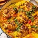 A close up of creamy saffron chicken with mascarpone and lemony cilantro dressing in a skillet with a serving spoon. Next to this is a stack of 2 plates and a bowl of the lemony cilantro dressing.