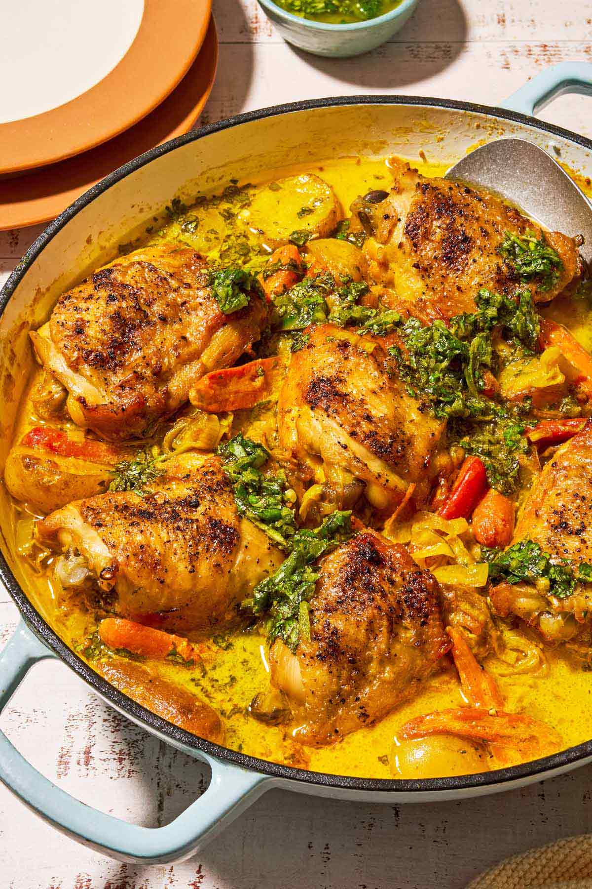 A close up of creamy saffron chicken with mascarpone and lemony cilantro dressing in a skillet with a serving spoon. Next to this is a stack of 2 plates and a bowl of the lemony cilantro dressing.