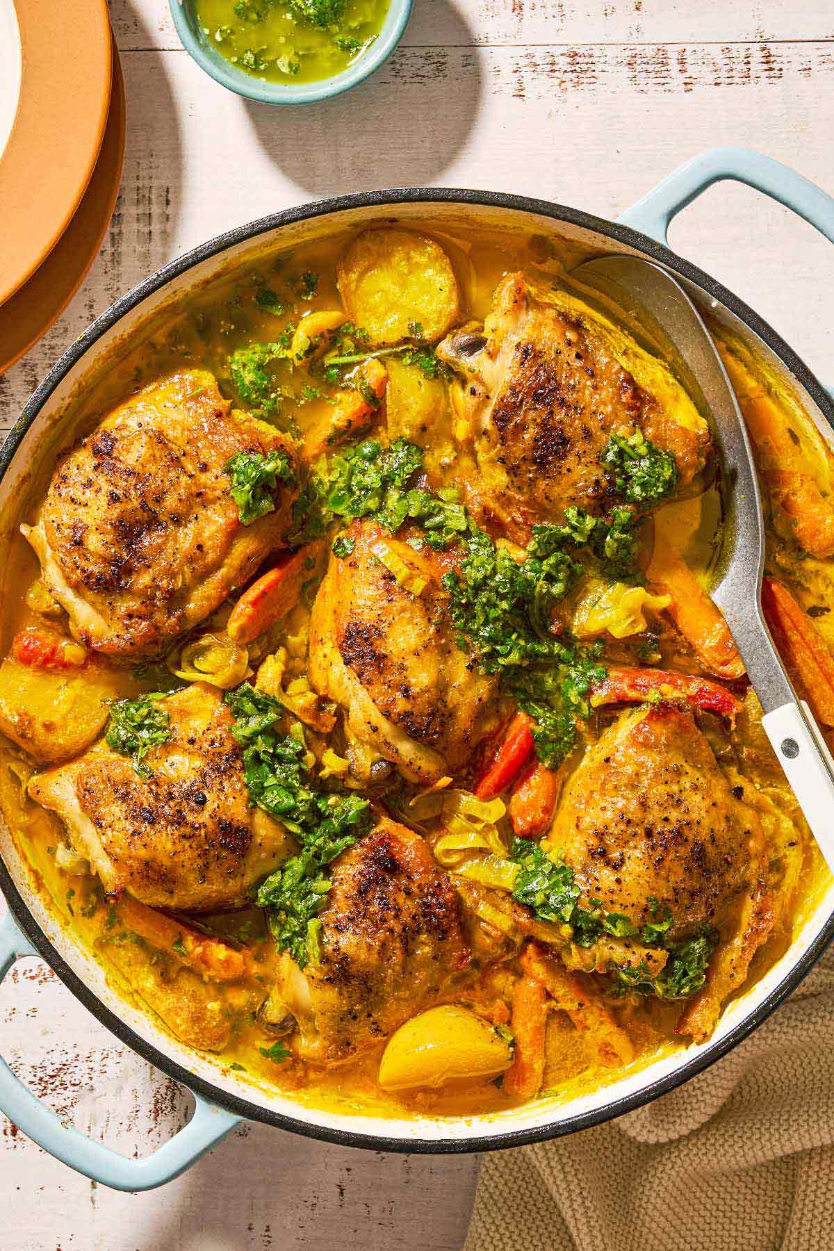 An overhead photo of creamy saffron chicken with mascarpone and lemony cilantro dressing in a skillet with a serving spoon. Next to this is a stack of two plates and a small bowl of the lemony cilantro dressing.