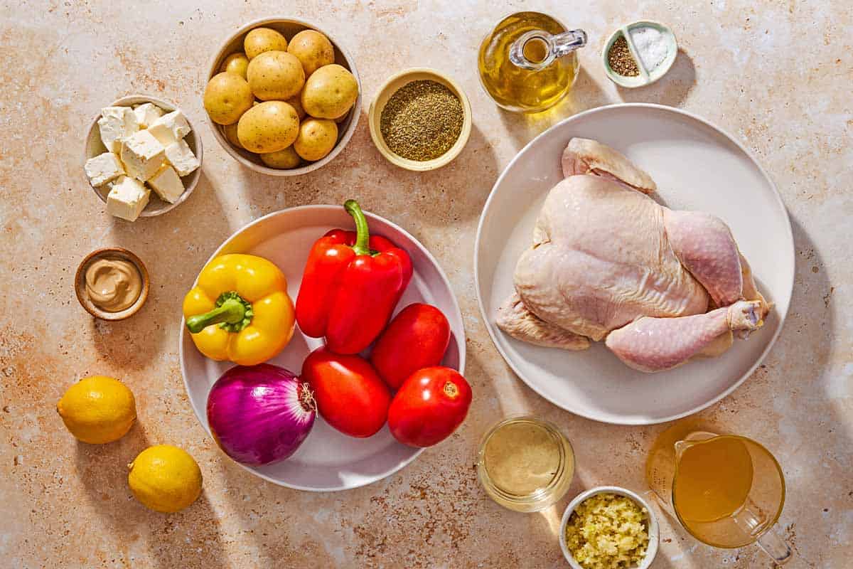 Ingredients for oven roasted whole chicken and potatoes including a whole chicken italian seasoning, garlic, dijon mustard, lemons, olive oil, salt, black pepper, baby potatoes, roma tomatoes, bell peppers, red onion, chicken stock, white wine, and feta cheese.