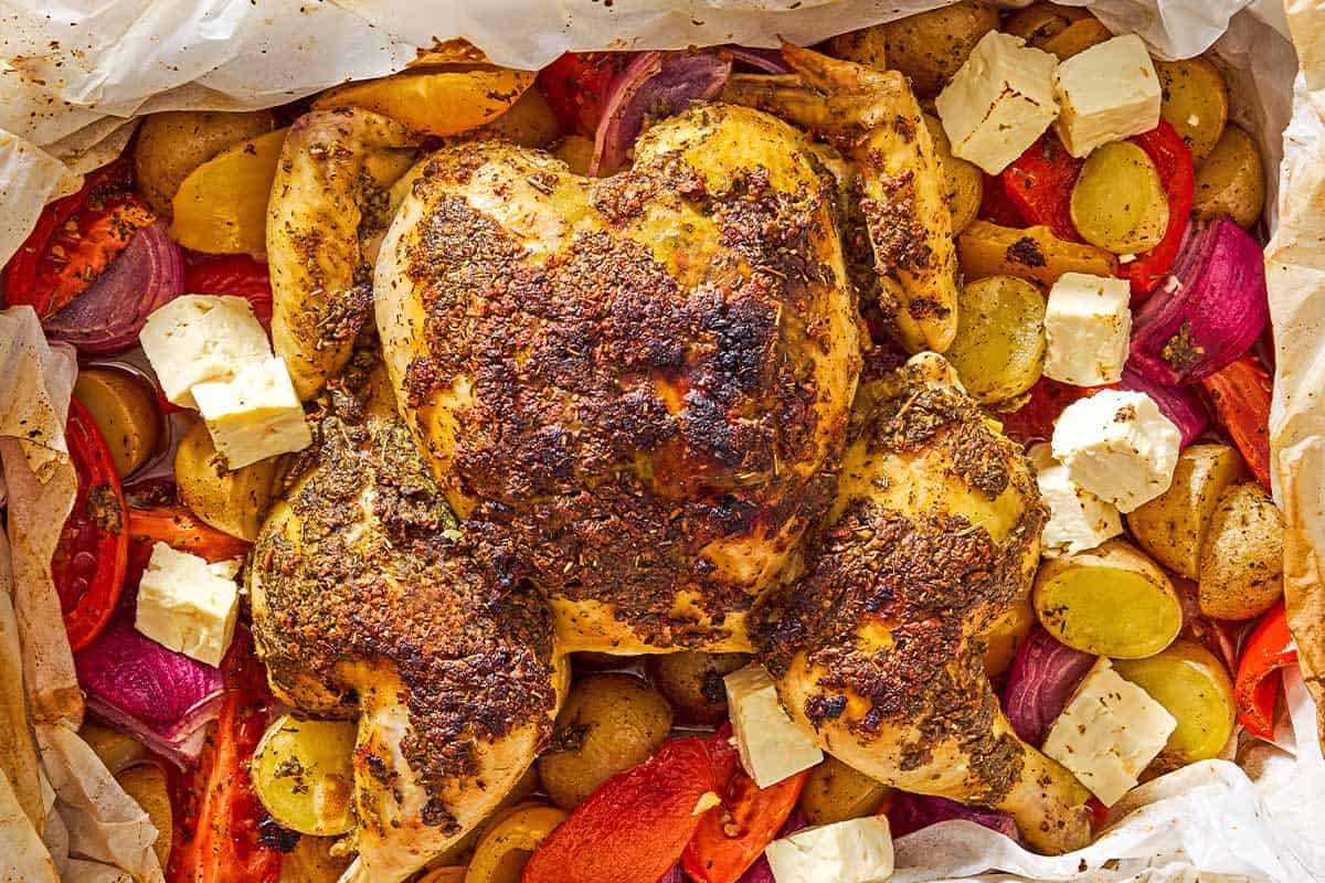 An overhead photo of oven roasted whole chicken and potatoes along with other vegetables and cubed feta in a parchment-line roasting pan.