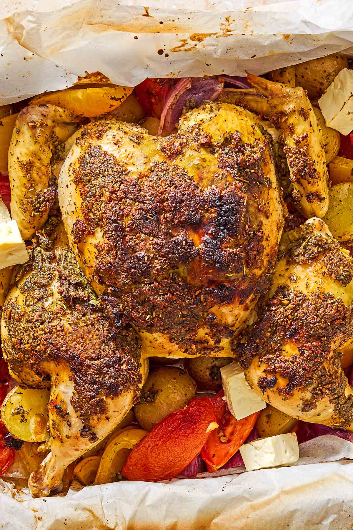 An overhead close up photo of oven roasted whole chicken and potatoes along with other vegetables and cubed feta in a parchment-line roasting pan.