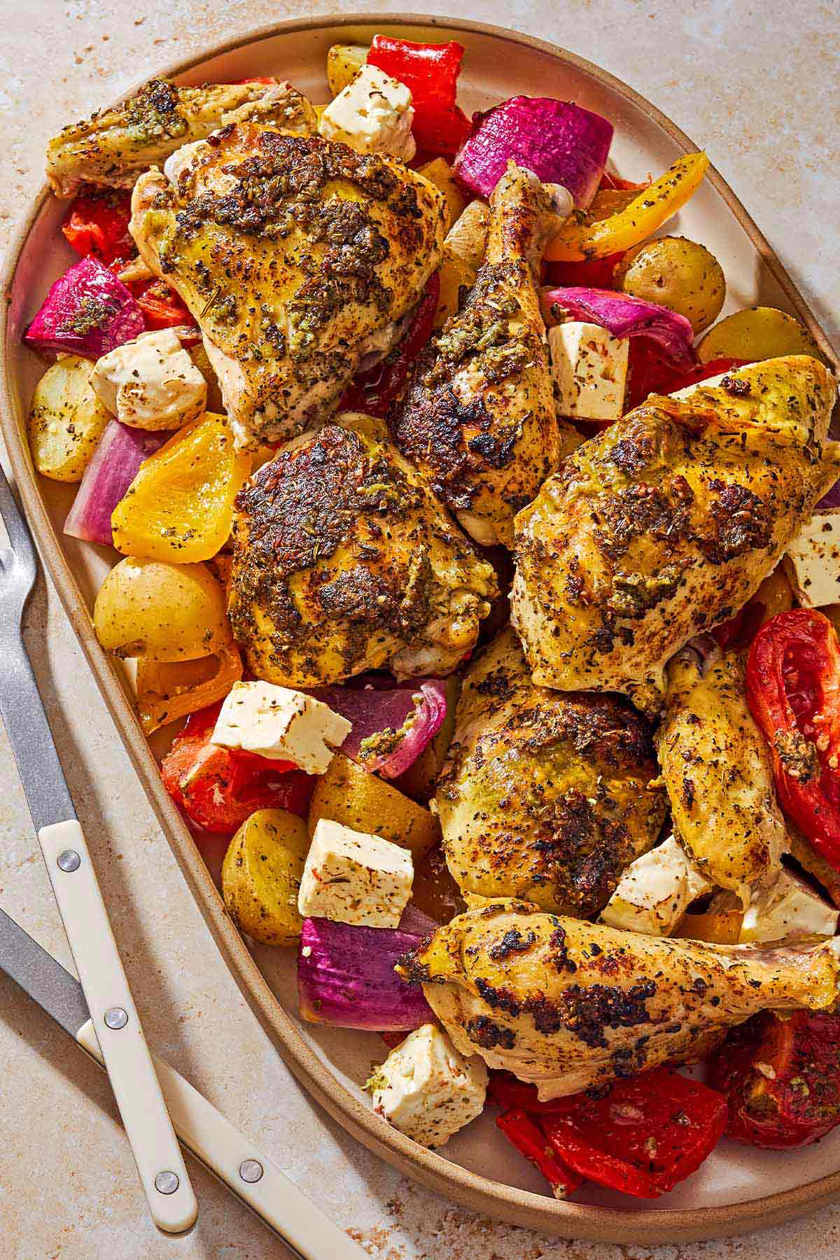 An overhead photo of oven roasted whole chicken and potatoes along with other vegetables and cubed feta on a platter next to serving utensils.