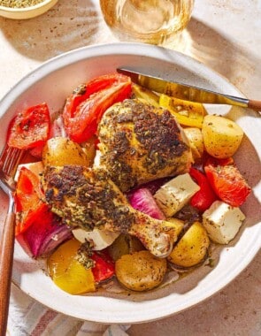 An overhead photo of a serving of oven roasted whole chicken and potatoes along with other vegetables and cubed feta on plate with a knife and fork. Next to this is a cloth napkin, a glass of white wine, and a bowl of Italian seasoning.