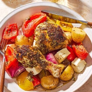 An overhead photo of a serving of oven roasted whole chicken and potatoes along with other vegetables and cubed feta on plate with a knife and fork. Next to this is a cloth napkin, a glass of white wine, and a bowl of Italian seasoning.