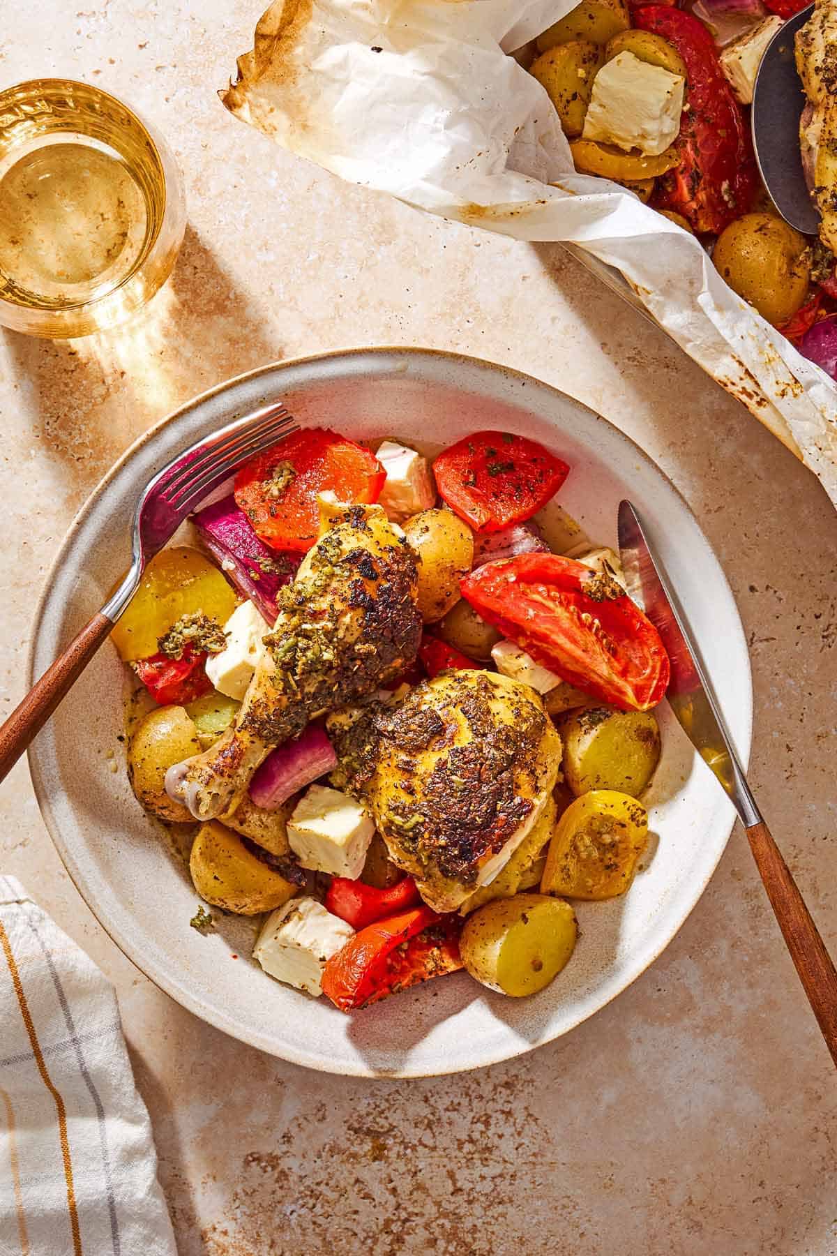 An overhead photo of a serving of oven roasted whole chicken and potatoes along with other vegetables and cubed feta on plate with a knife and fork. Next to this is a cloth napkin, a glass of white wine, and the rest of the chicken and vegetables in a roasting pan.