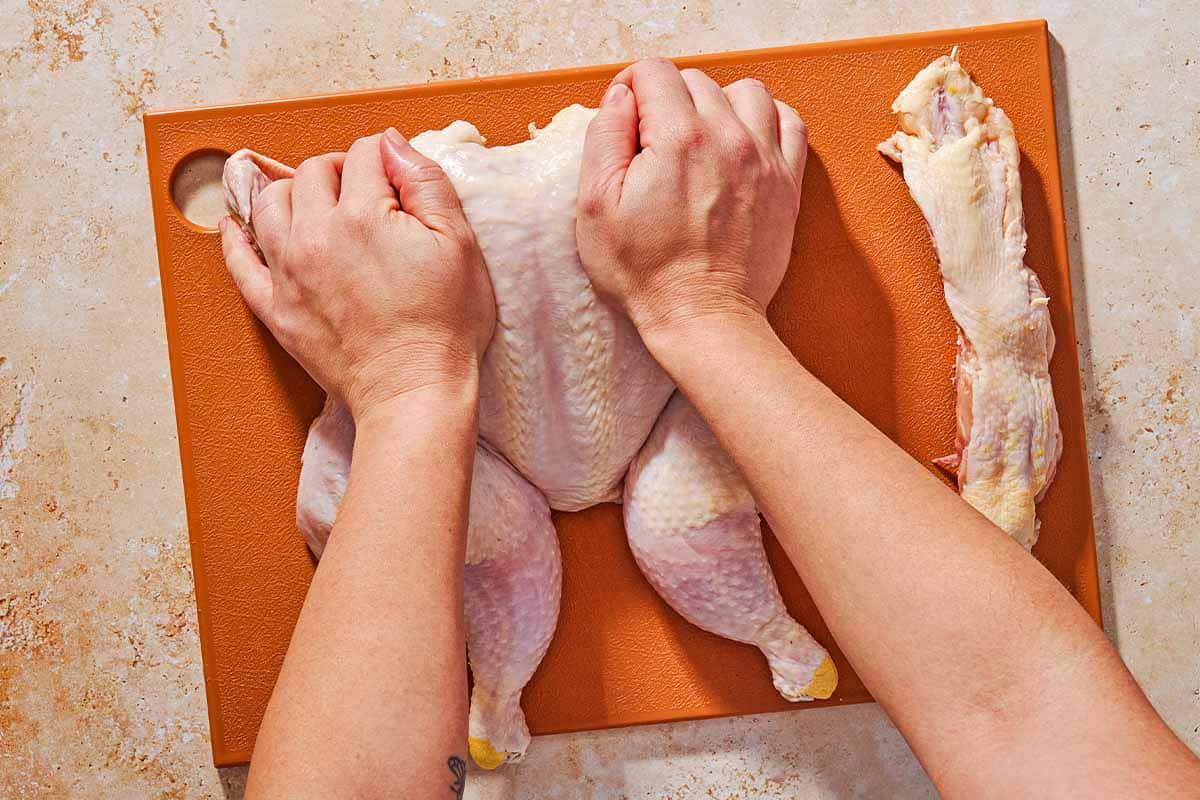 An overhead photo of a whole raw chicken being spatchcocked on a cutting board. Next to this is the removed backbone.