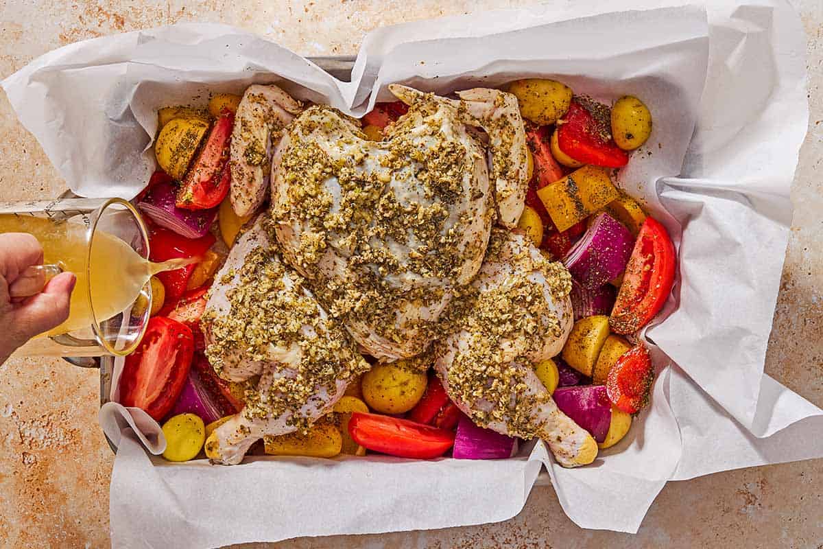 An overhead photo of chicken broth being poured into a parchment-line roasting pan to the side of the whole uncooked chicken vegetables.