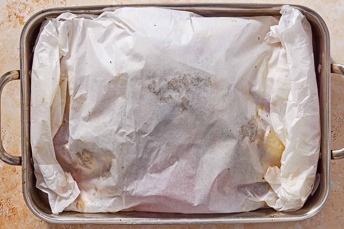 An overhead photo of oven roasted whole chicken and potatoes along with other vegetables and cubed feta completely enveloped in parchment paper in a roasting pan.