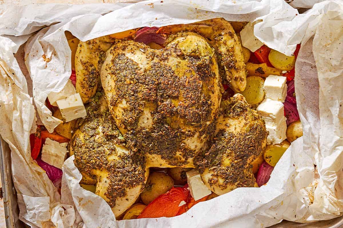 An overhead photo of oven roasted whole chicken and potatoes along with other vegetables and cubed feta in a parchment-line roasting pan.