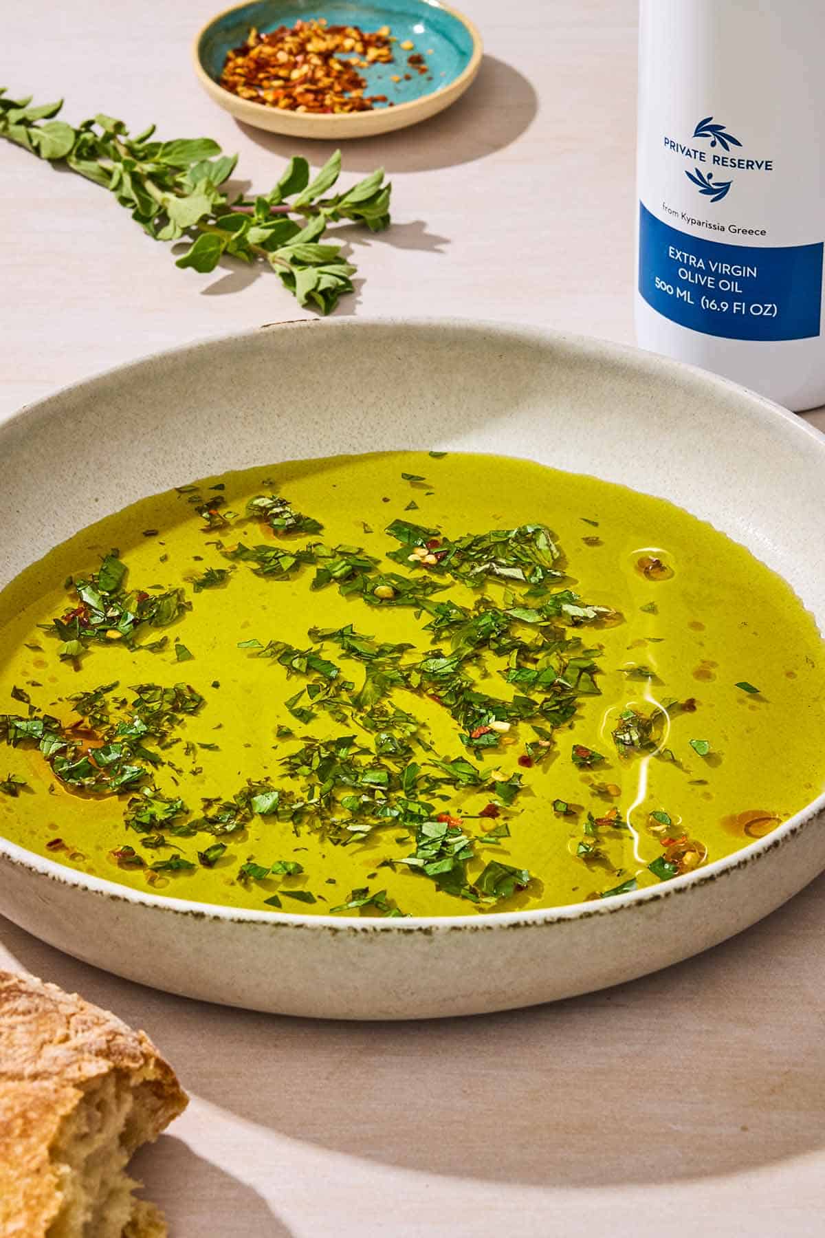 A bowl of olive oil topped with fresh herbs and red pepper flakes. Next to this is a bowl of red pepper flakes, a sprig of fresh herbs, a piece of crusty bread, and a bottle of olive oil.