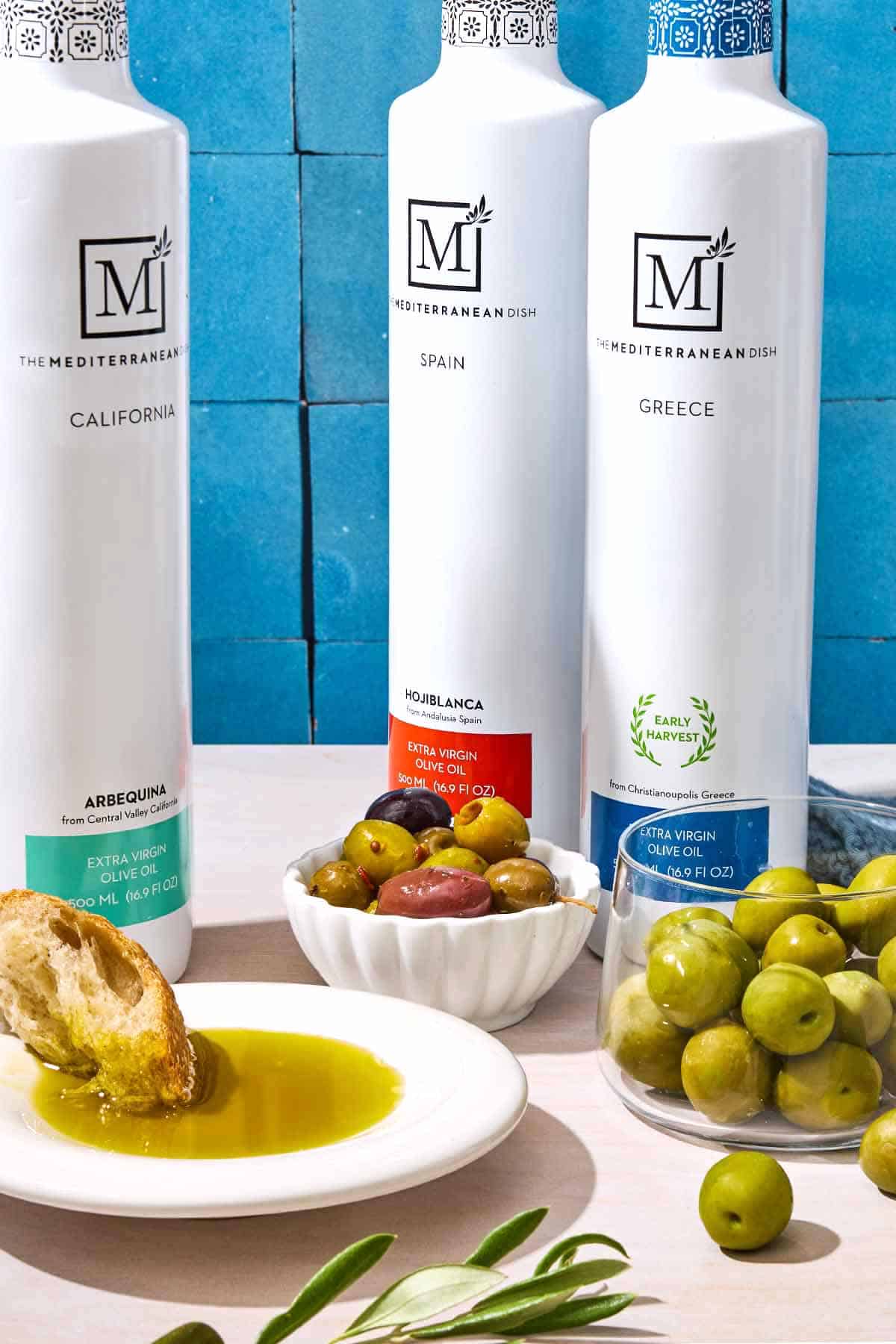 3 bottles of olive oil from The Mediterranean Dish surrounded by bowls of olives and a plate with some olive oil and piece of crusty bread.