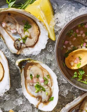 A close up of 3 raw oysters with mignonette sauce on a bed of ice on a tray with lemon wedges and a bowl with the rest of the sauce and a spoon.