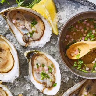 A close up of 3 raw oysters with mignonette sauce on a bed of ice on a tray with lemon wedges and a bowl with the rest of the sauce and a spoon.