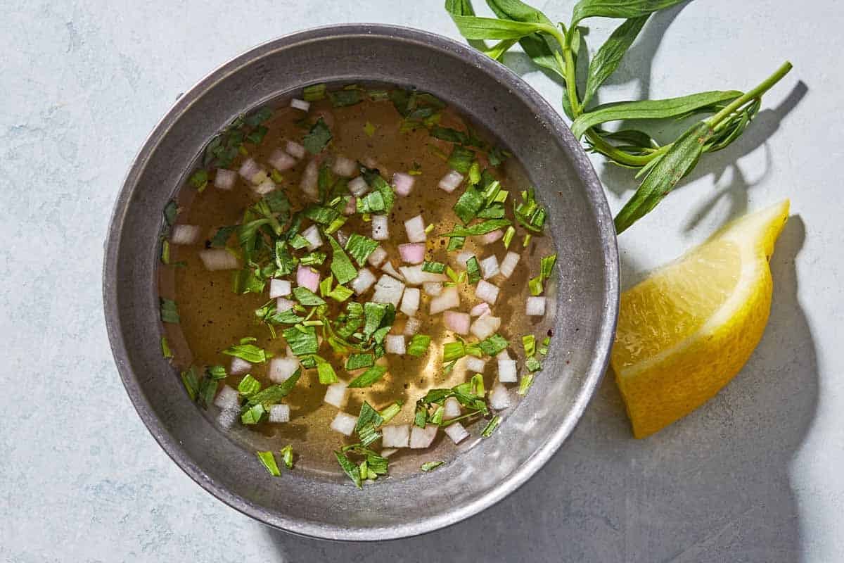 An overhead photo of mignonette sauce in a bowl next to fresh herbs and a lemon wedge.