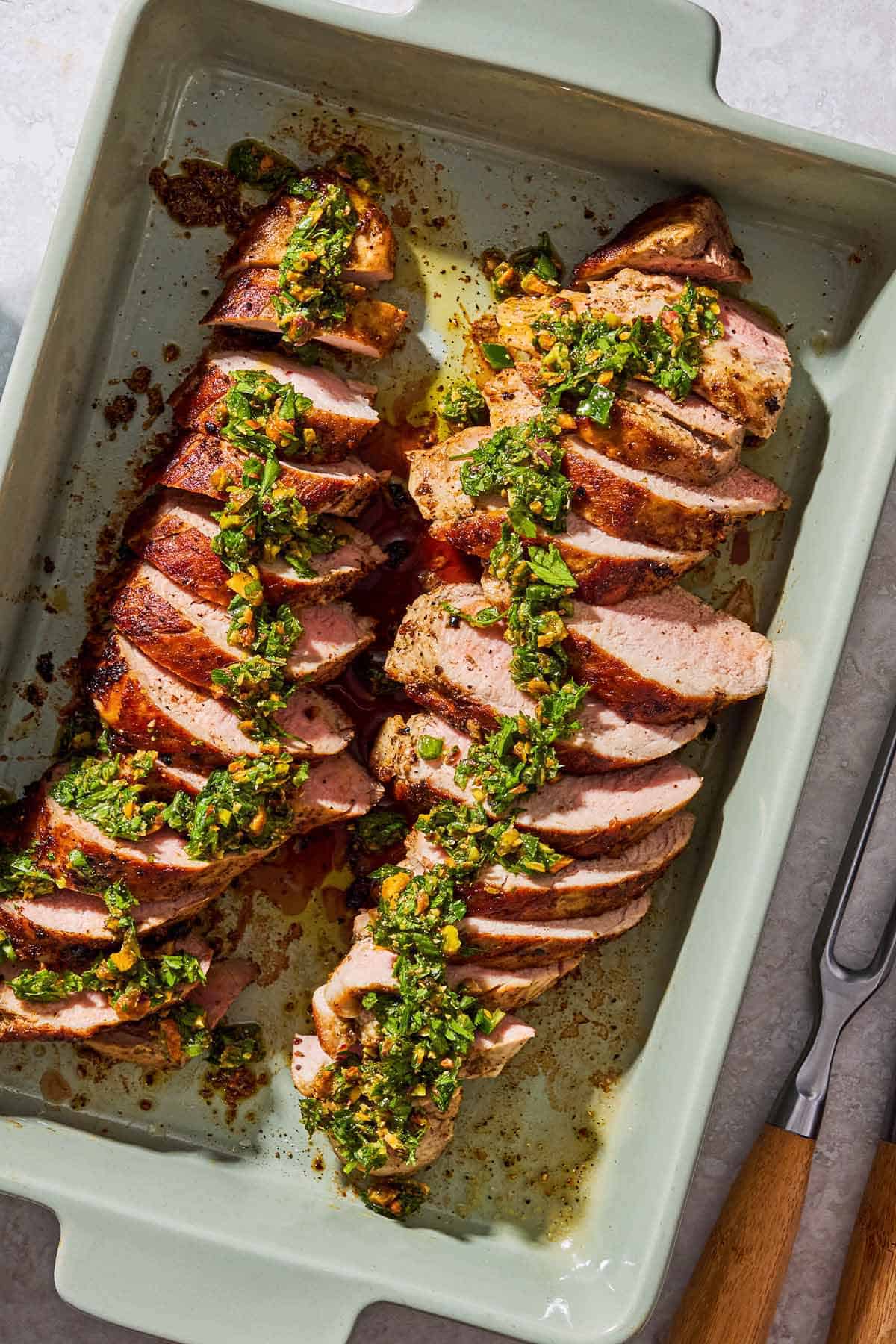 Sliced baked pork tenderloin topped with herb and pistachio salsa in a baking dish.