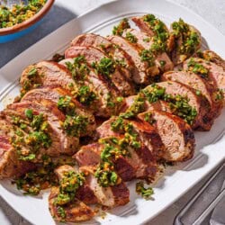 Sliced baked pork tenderloin topped with herb and pistachio salsa on a platter. Next to this is a chef's knife and a serving fork, and a bowl with the rest of the salsa.