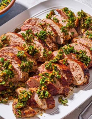 Sliced baked pork tenderloin topped with herb and pistachio salsa on a platter. Next to this is a chef's knife and a serving fork, and a bowl with the rest of the salsa.