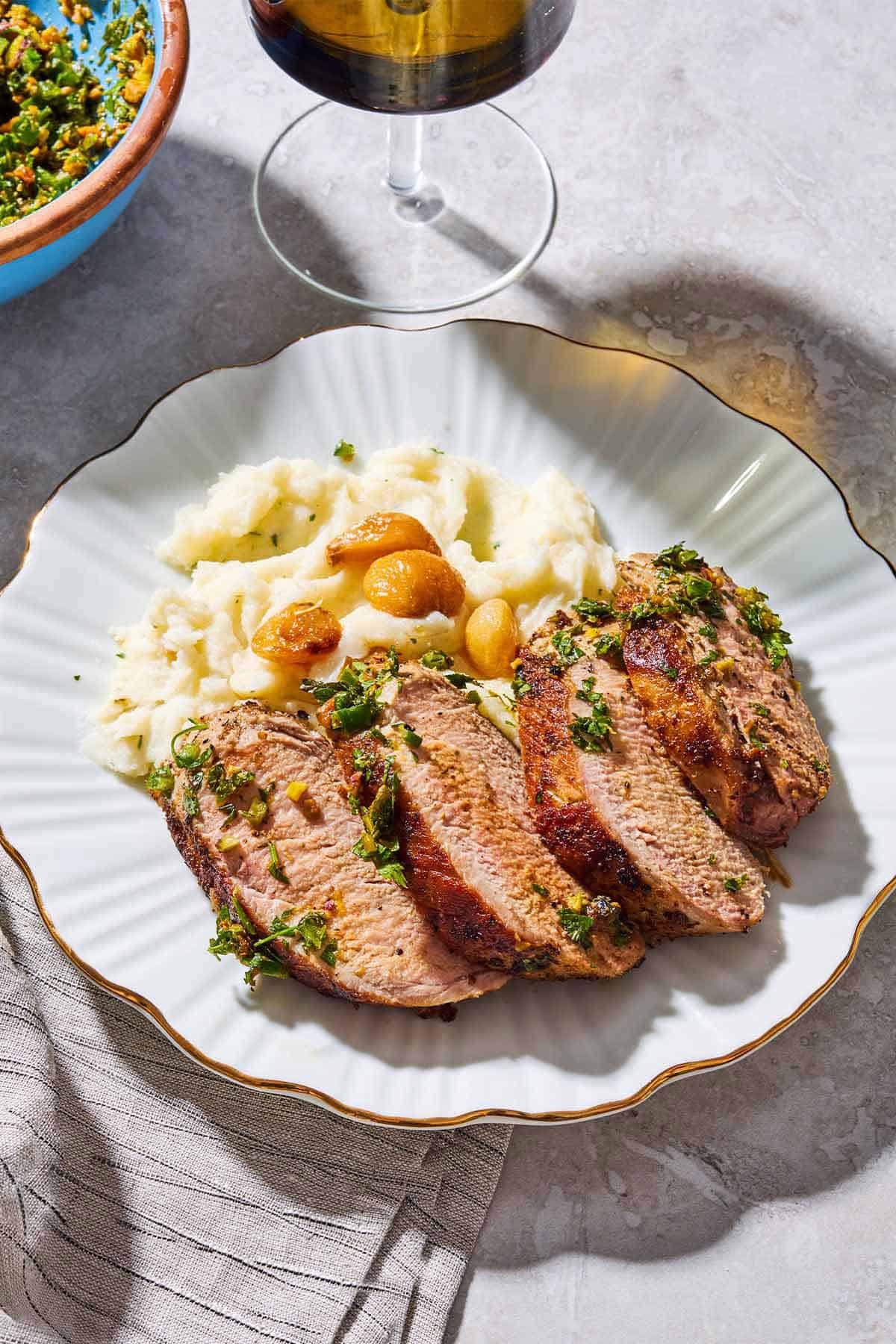 Sliced baked pork tenderloin topped with herb and pistachio salsa on a plate with garlic mashed potatoes. Next to this is a cloth napkin, a bowl of the salsa, and a glass of wine.