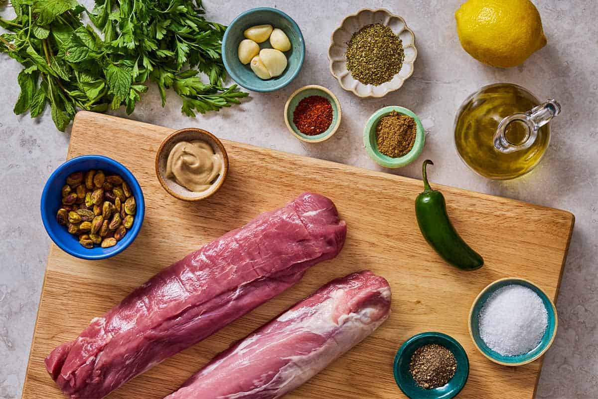 Ingredients for baked pork tenderloin including pork tenderloins, olive oil, garlic, dijon, oregano, coriander, aleppo pepper, jalapeno, parsley, mint, pistachios, lemon, salt and black pepper.