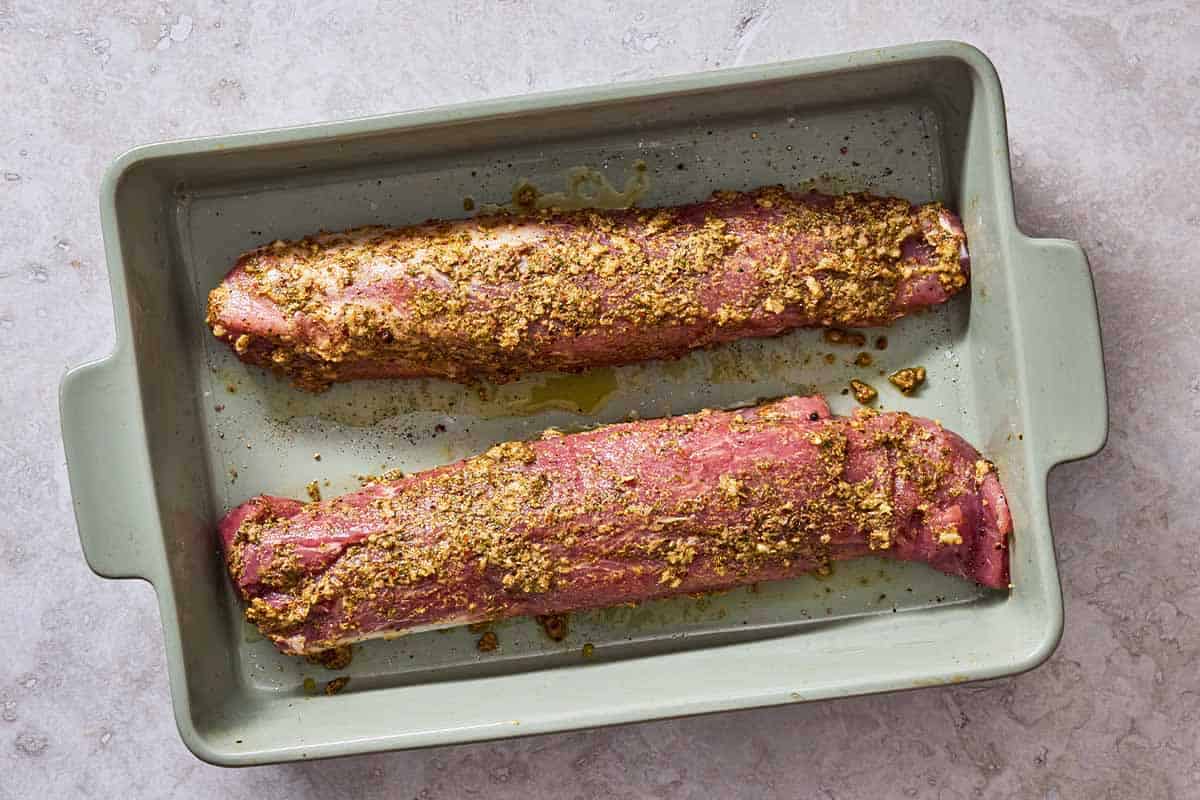 Two uncooked pork tenderloins being marinaded in a baking dish.