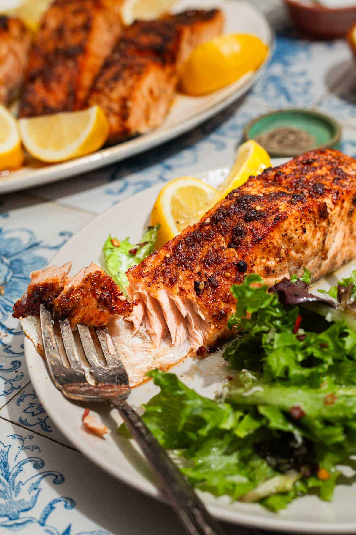 A close up of an air fried salmon fillet on a plate with lemon wedges, salad and a fork. In the background in is the rest of the salmon on a platter and a bowl of black pepper.