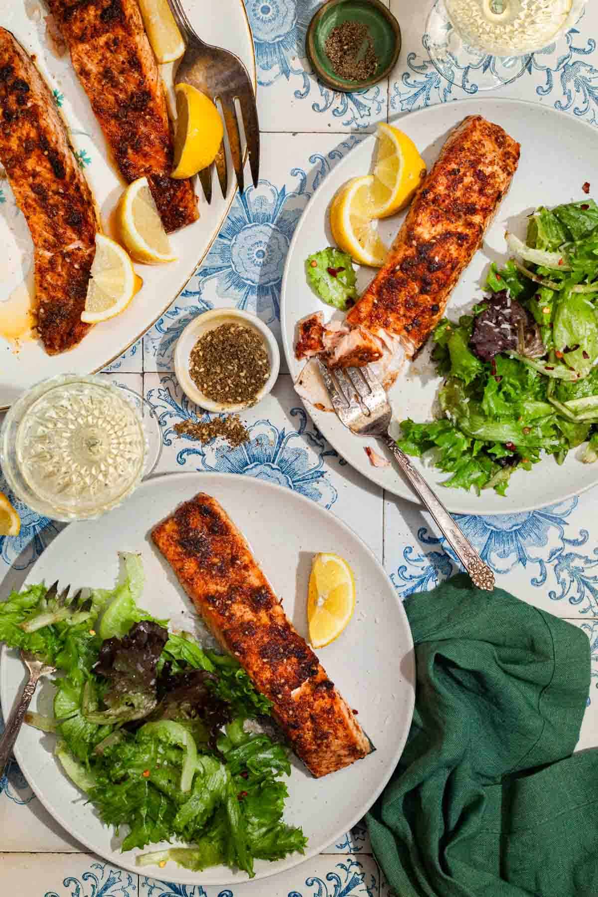 An overhead photo of 2 plates with air fryer salmon, salad, lemon wedges and forks. Next to these is a cloth napkin, 2 glasses of white wine, bowls of black pepper and za'atar, and the rest of the salmon on a serving platter with a fork.