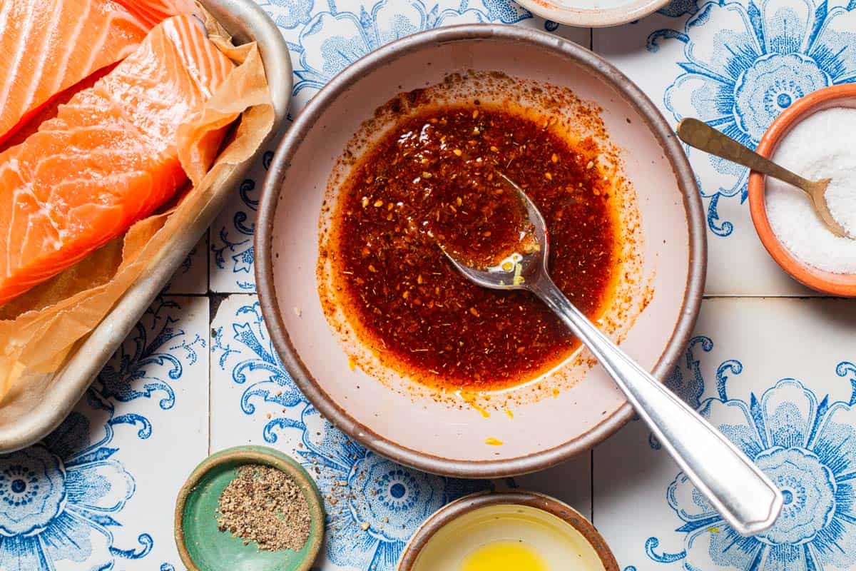 An overhead photo of the marinade for the air fryer salmon in a bowl with a spoon, Next to this is a parchment lined sheet pan with uncooked salmon and bowls of salt and black pepper.