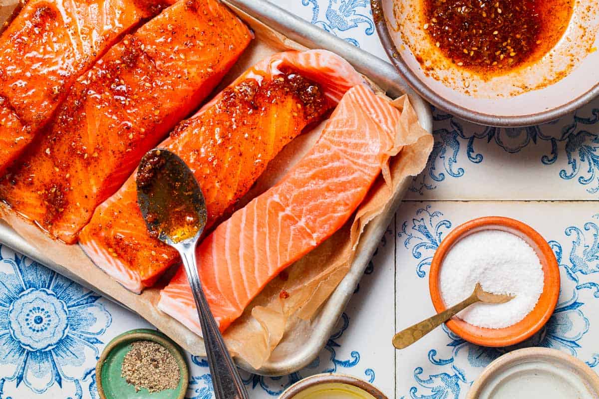 An overhead photo of 4 uncooked salmon fillets on a parchment lined sheet pan with a spoon, three are coated in the marinade. Next to this is the rest of the marinade in a bowl with a spoon, and bowls of salt and black pepper.