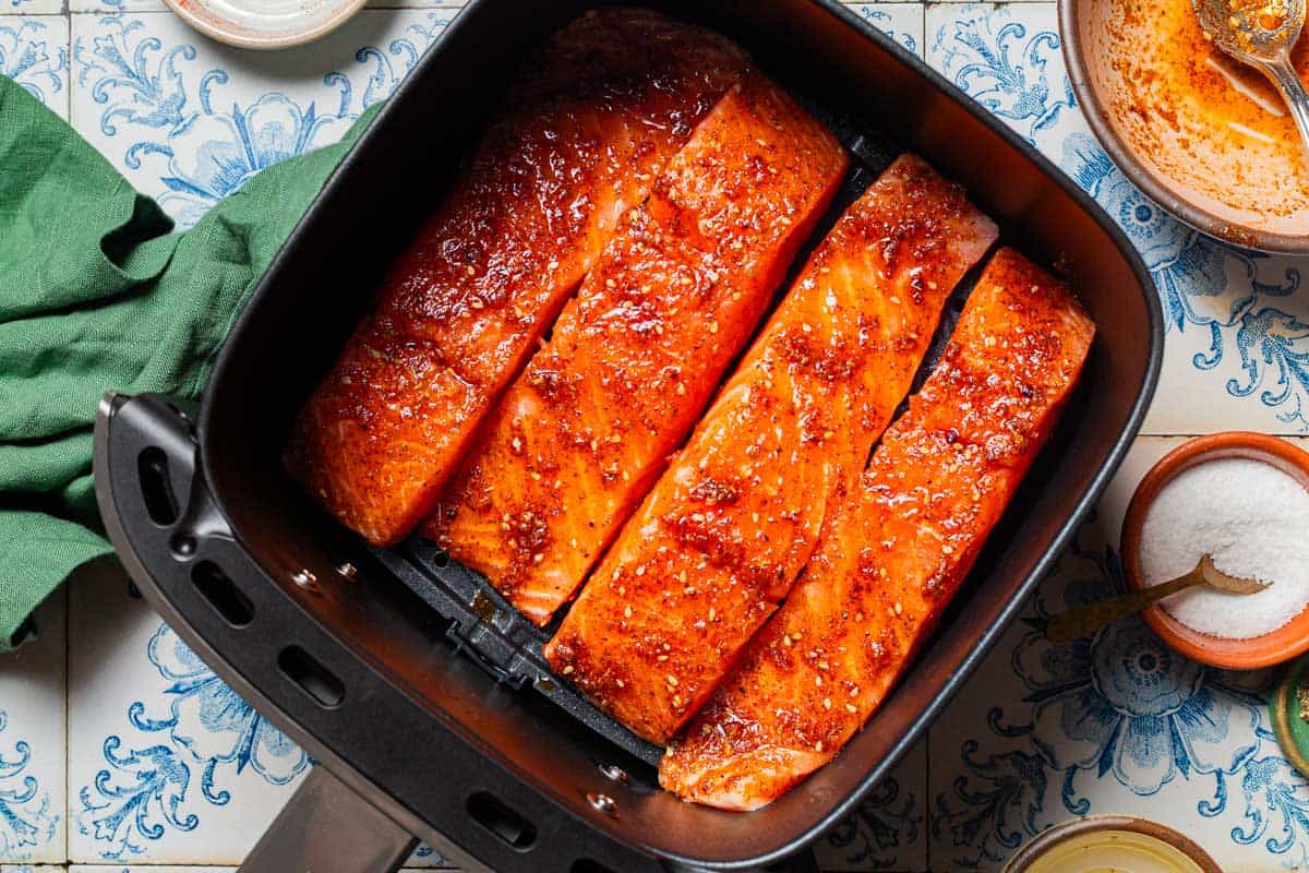 An overhead photo of 4 uncooked marinaded salmon fillets in the basket of an air fryer. Next to this is a cloth napkin, an empty bowl with a spoon, and a bowl of salt.