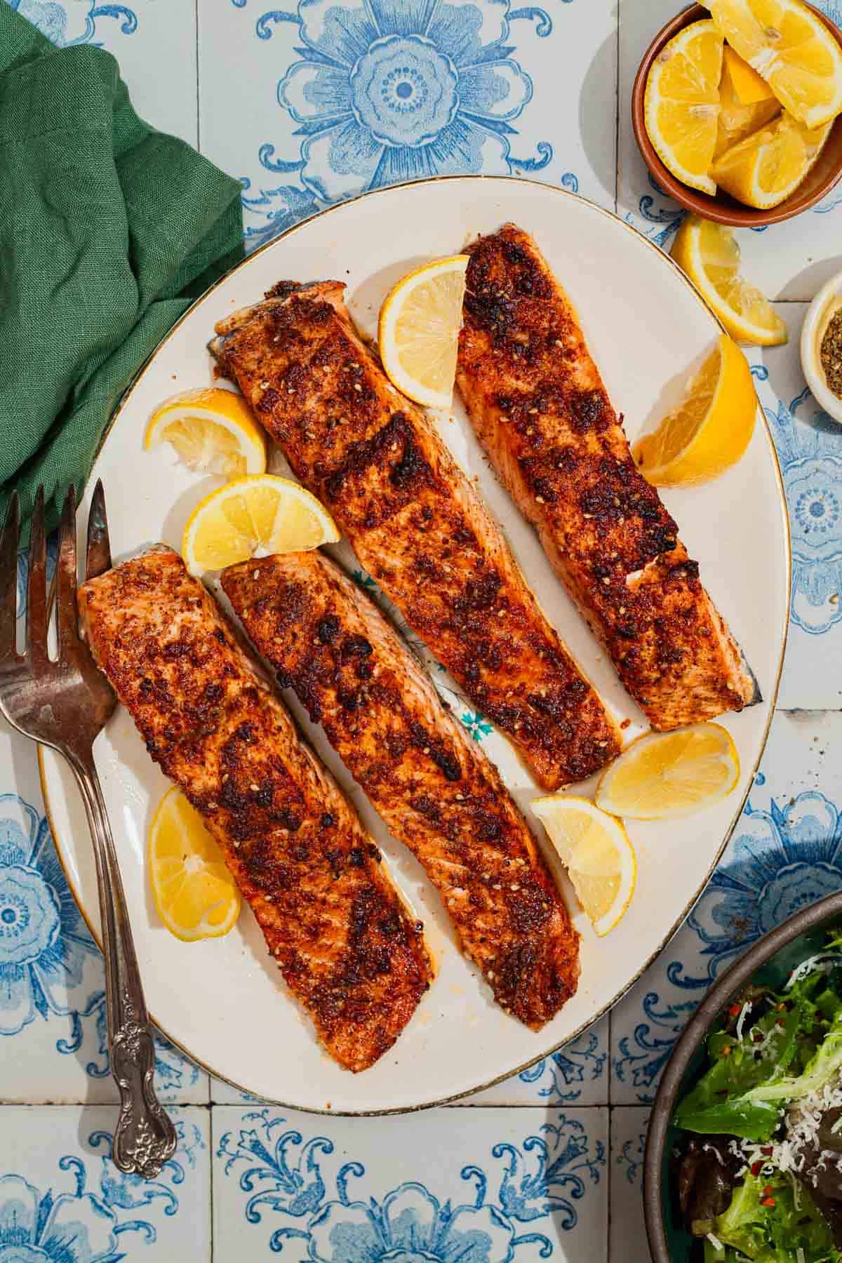 An overhead photo of 4 air fried salmon fillets on a platter with a fork and some lemon wedges. Next to this is a cloth napkin and a bowl of lemon wedges.