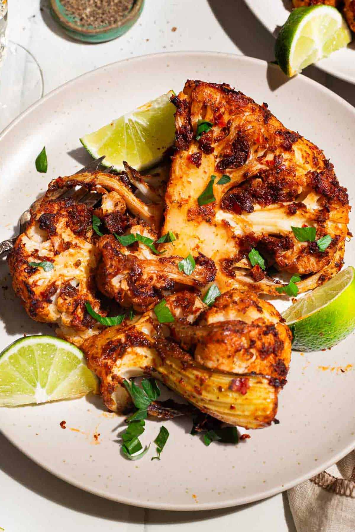 A close up photo of a serving of spicy baked cauliflower topped with chopped parsley and lime wedges on a plate.