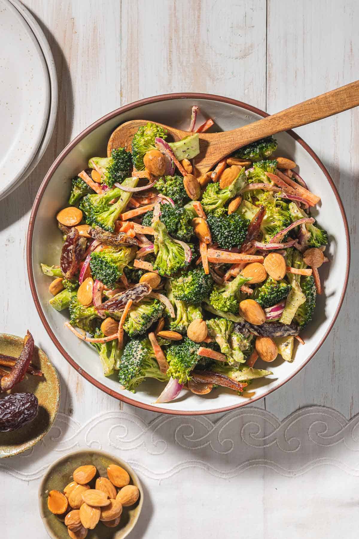 An overhead photo of broccoli salad in a serving bowl with a wooden spoon. Next to this is a stack of 2 plates and small bowls of dates and toasted almonds.