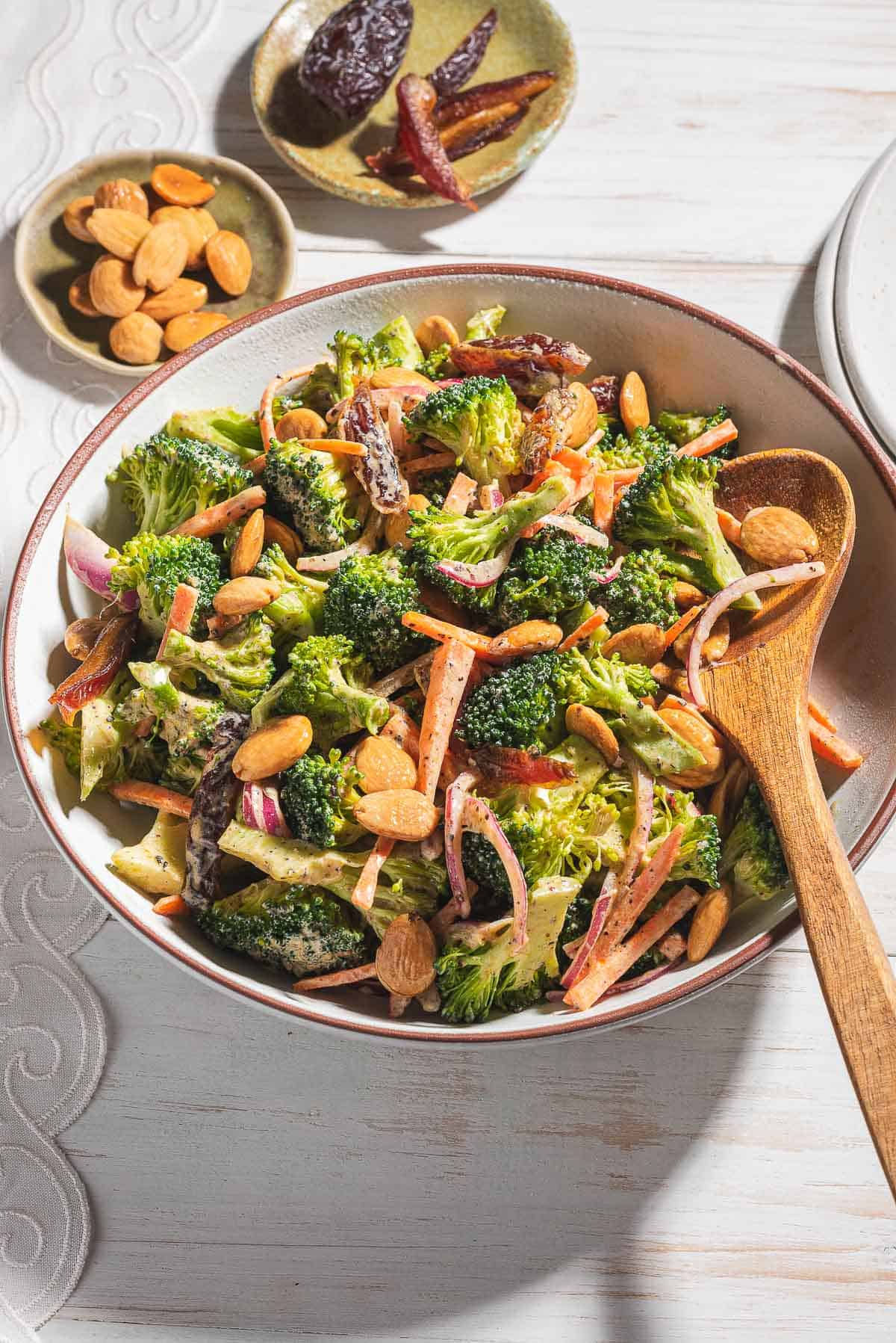 A photo of broccoli salad in a serving bowl with a wooden spoon. Next to this is a stack of 2 plates and small bowls of dates and toasted almonds.