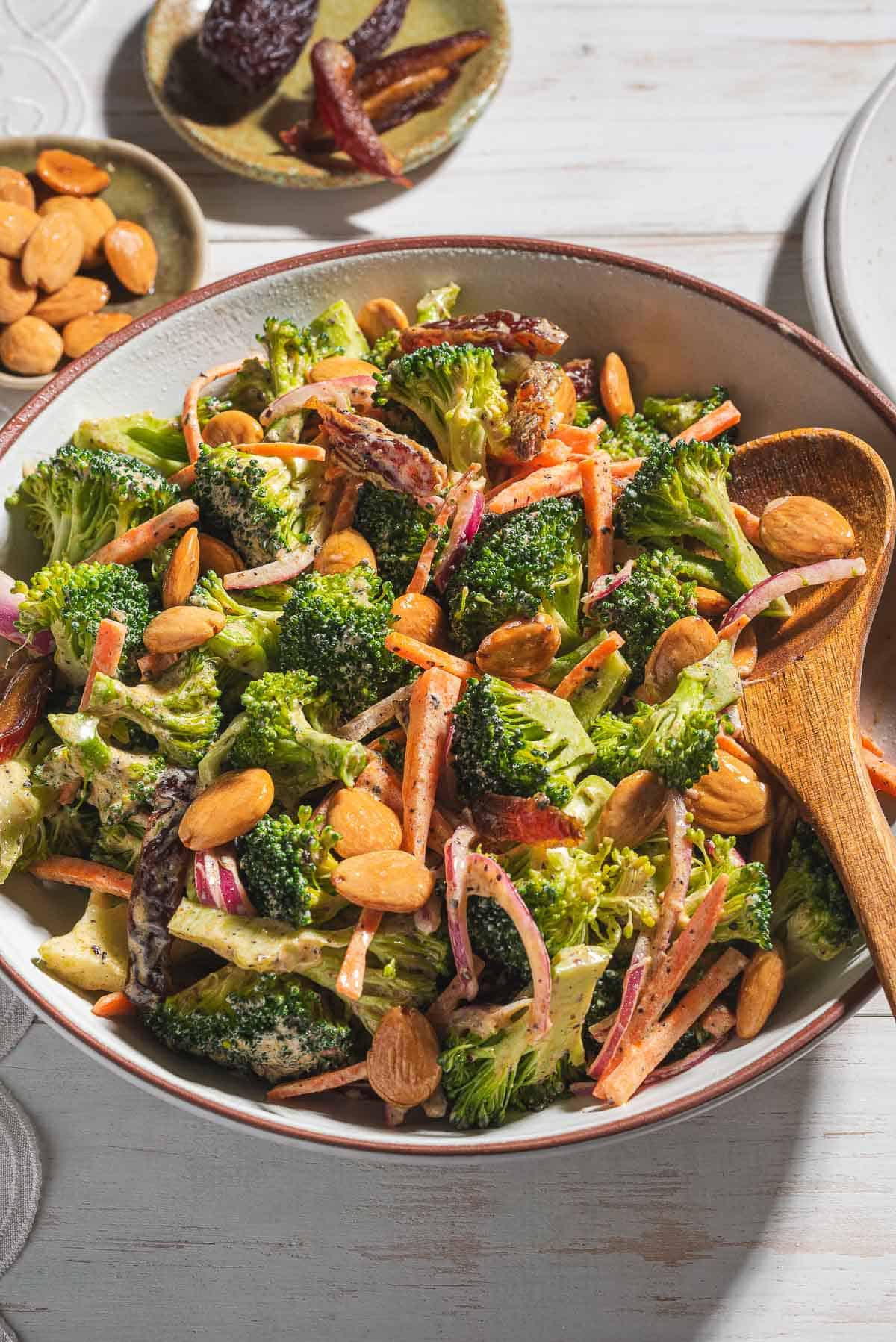 A close up of broccoli salad in a serving bowl with a wooden spoon. Next to this is a stack of 2 plates and small bowls of dates and toasted almonds.