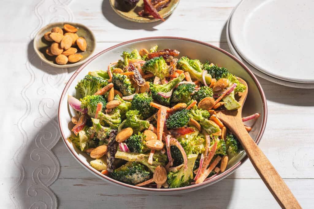 A photo of broccoli salad in a serving bowl with a wooden spoon. Next to this is a stack of 2 plates and small bowls of dates and toasted almonds.