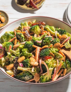 A photo of broccoli salad in a serving bowl with a wooden spoon. Next to this is a stack of 2 plates and small bowls of dates and toasted almonds.